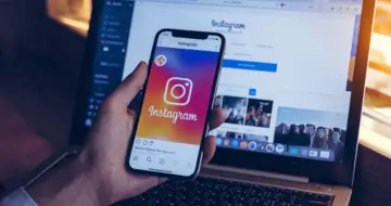 Pessoa usando o aplicativo Instagram no smartphone em frente a um laptop, representando estratégias para anunciar no Instagram e alcançar maior visibilidade na rede social.